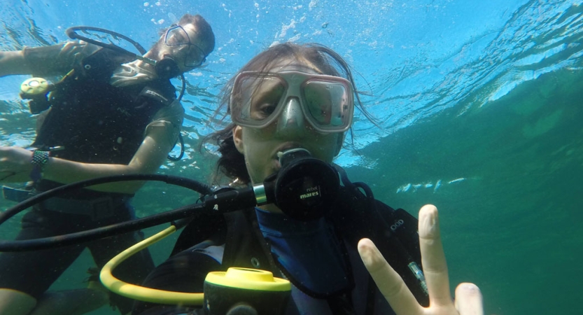 Two people are scuba diving underwater and look at the camera. The person in the foreground gives the camera a peace sign. 